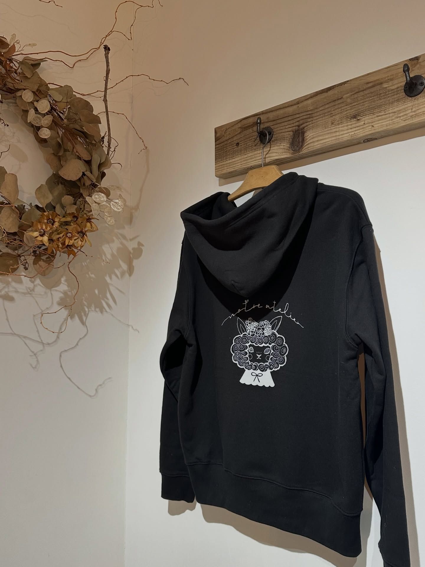 壁のフックに掛かった、ひつじのロゴ入りの黒いオリジナルパーカー